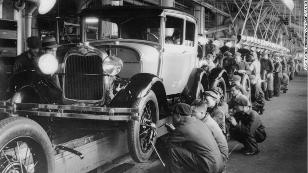 Black-and-white photo of workers on an early automobile assembly line, illustrating specialized engineering teams