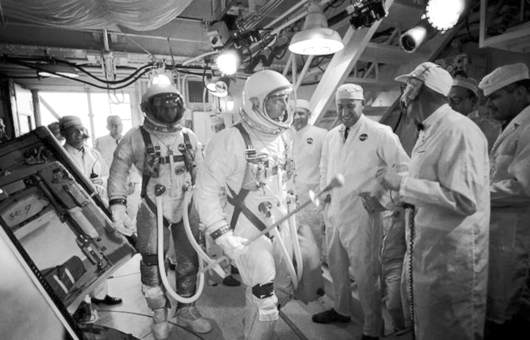 Black-and-white photo of astronauts in pressure suits with technicians before a capsule, from the space program era
