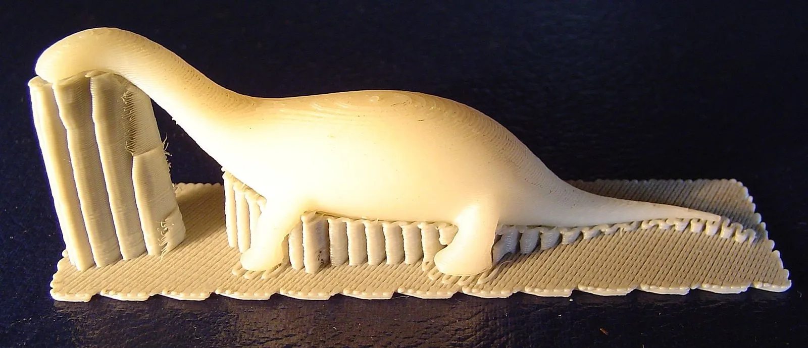 FDM print of a dinosaur figure with visible layer lines and supports on a raft, showing plastic applied layer by layer