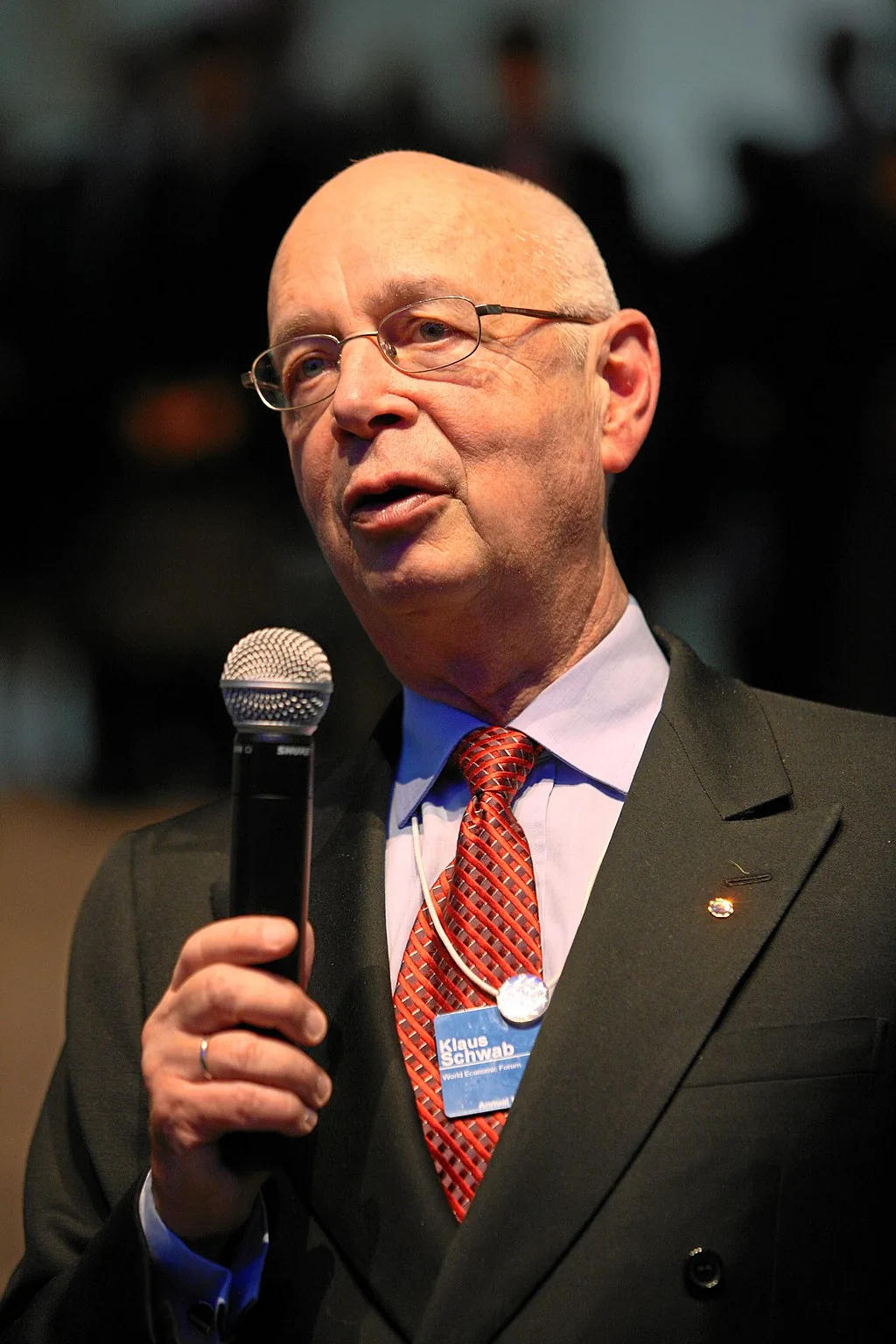 Klaus Schwab speaking into a microphone at the World Economic Forum, wearing his WEF name badge